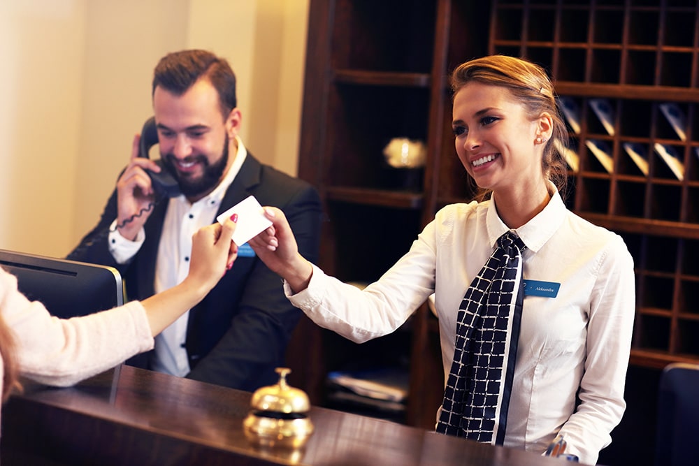 Smiling hotel receptionist providing a key card to a guest, while another staff member takes a call, illustrating the excellent guest experience enabled by reliable hospitality IT support.